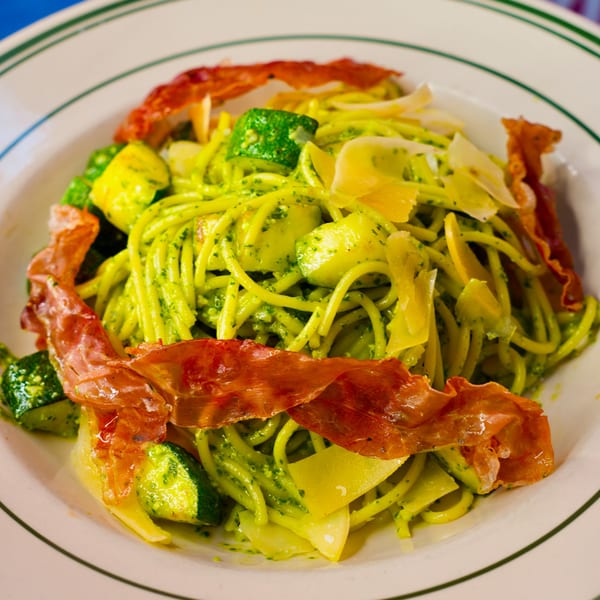 Green pesto pasta with zucchini, shaved cheese, and crispy prosciutto in a white bowl.